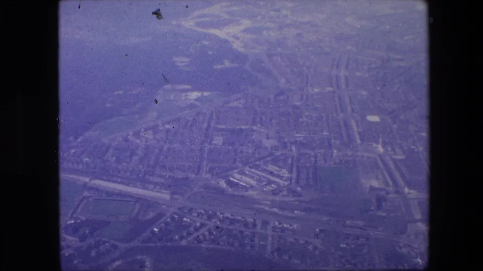 1960s 8mm-style aerial view from an airplane window showing a hazy urban grid below, with no identifiable landmark visible in the frame.