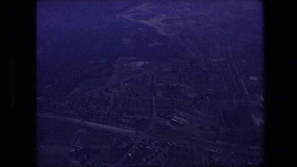 Underexposed purple-tinted aerial view of a dense cityscape below an aircraft, framed by black film edges and window shadow.