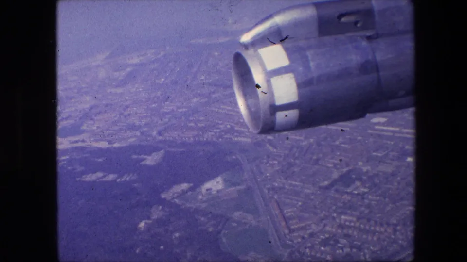 Passenger-window view with a jet engine nacelle prominent in the foreground and a hazy urban landscape visible far below.