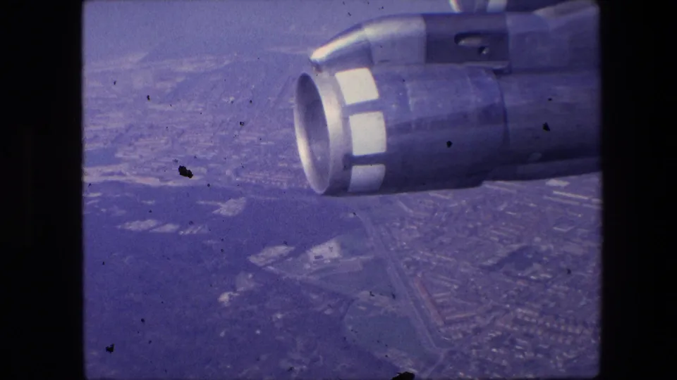 Vintage 8mm passenger-flight view showing the aircraft engine at right and a soft, purple-tinted city grid beneath the plane.