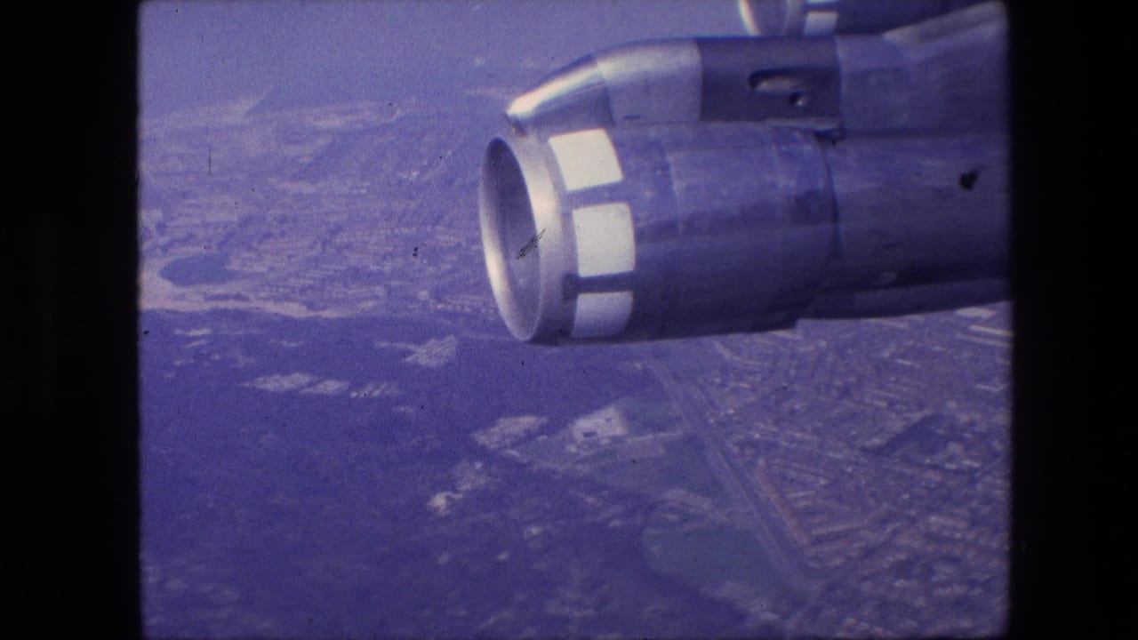 1967 8mm frame showing a jet engine nacelle outside an aircraft window with a hazy aerial view of roads and developed land below.