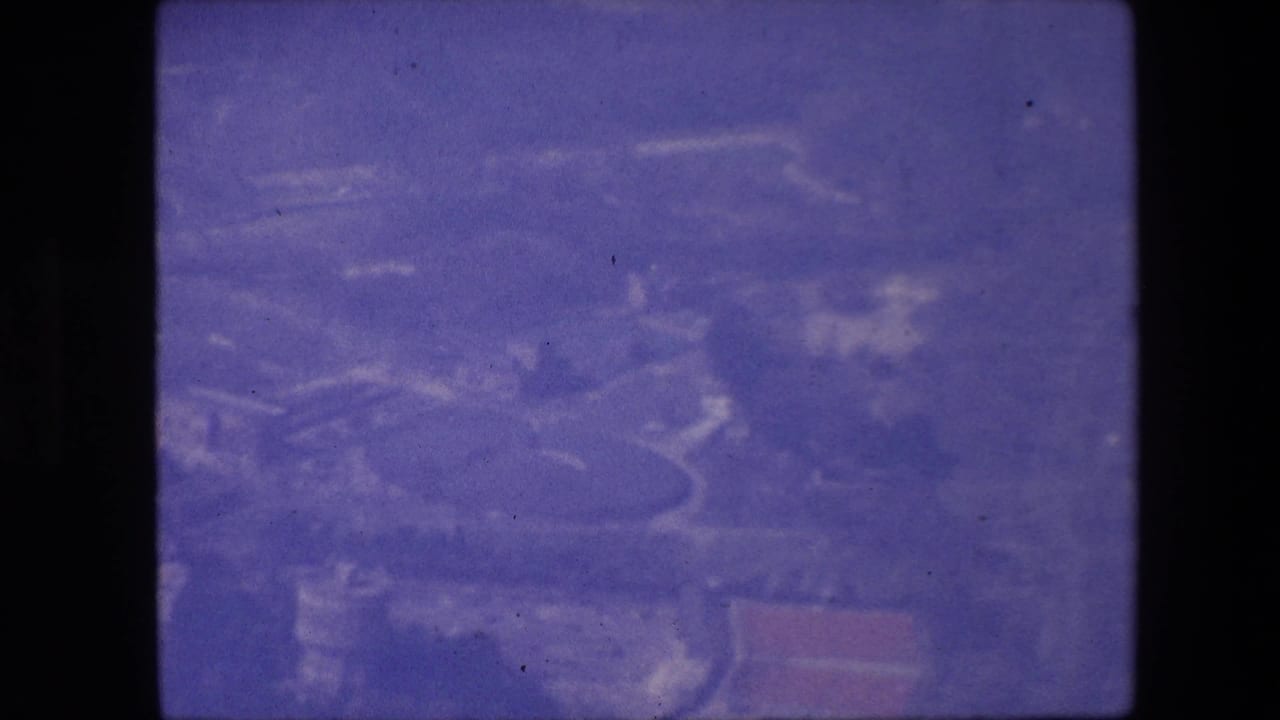 Purple-tinted 8mm aerial frame looking down at indistinct urban or suburban ground patterns, with no identifiable landmarks visible.