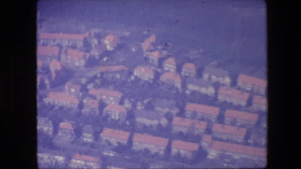 Aerial 8mm frame showing red-roofed residential buildings, streets, and clustered development below the aircraft.