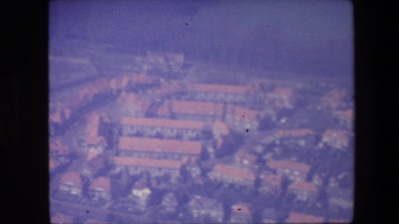 1967 8mm aerial view over a town area with rows of red-roofed buildings, streets, trees, and soft color fading.