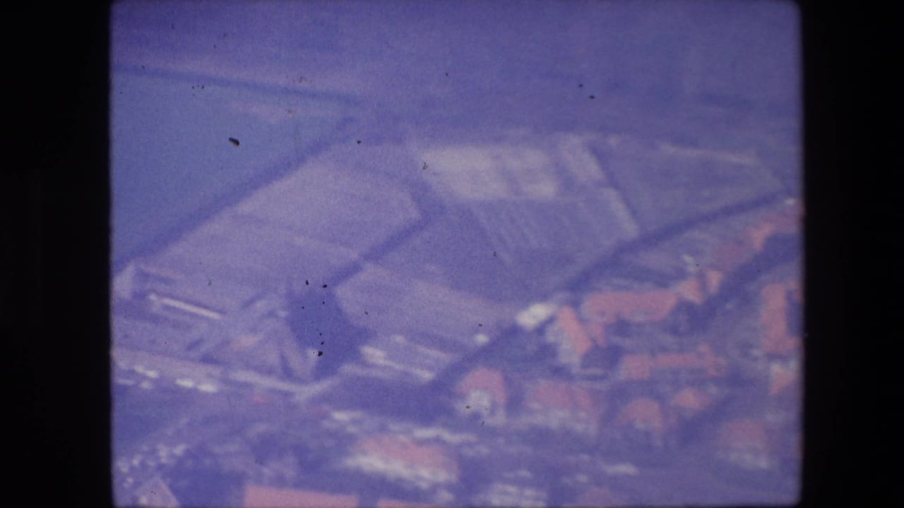 Aerial 8mm frame showing broad roof forms or airport-area structures with nearby red-roofed buildings, rendered with heavy softness and color shift.