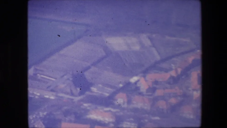 Aerial 8mm frame showing broad roof forms or airport-area structures with nearby red-roofed buildings, rendered with heavy softness and color shift.