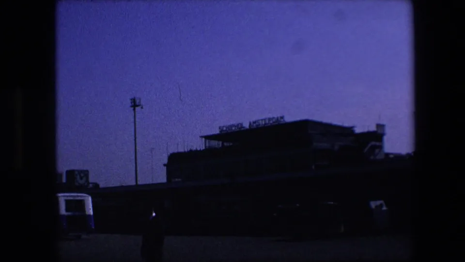 Underexposed 1967 8mm view of the Schiphol Amsterdam terminal exterior with a standing figure, terminal signage, pavement, and a vehicle edge visible.