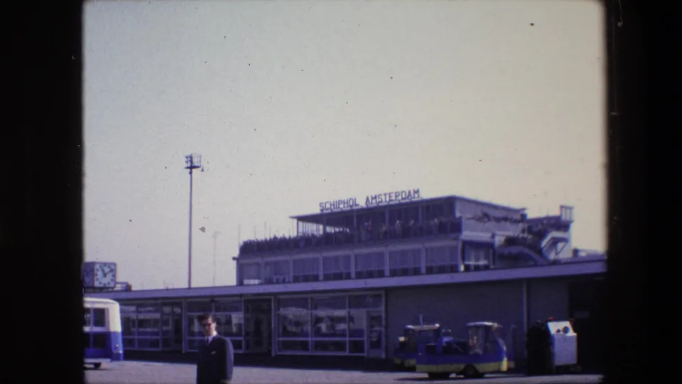 Clearer 1967 exterior view of Schiphol Amsterdam terminal showing a suited man in front of the building, a public clock, windowed facade, and small period vehicles.