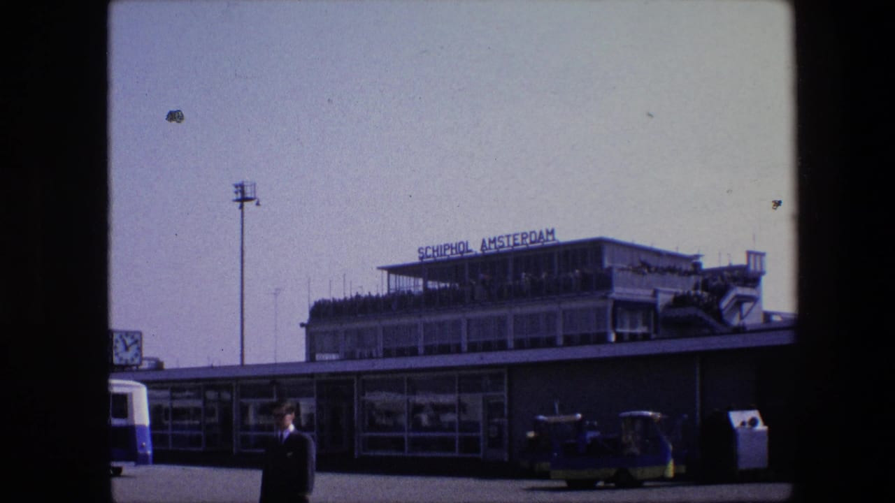 1967 8mm frame of the Schiphol Amsterdam terminal exterior, with the rooftop sign, observation deck crowd, light pole, bus edge, service vehicles, and a suited man waiting in front.