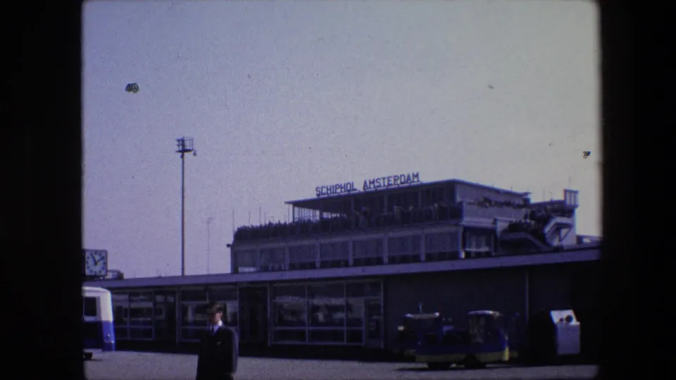 1967 8mm frame of the Schiphol Amsterdam terminal exterior, with the rooftop sign, observation deck crowd, light pole, bus edge, service vehicles, and a suited man waiting in front.