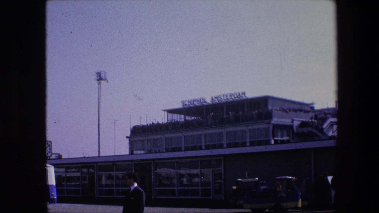 1967 8mm frame showing the Schiphol Amsterdam airport terminal from outside, with the sign on the roof, people on the upper deck, a suited man in front, and ground vehicles along the curb.