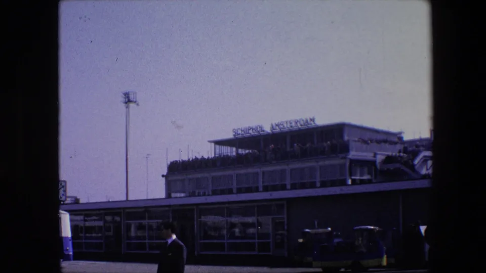 1967 8mm frame showing the Schiphol Amsterdam airport terminal from outside, with the sign on the roof, people on the upper deck, a suited man in front, and ground vehicles along the curb.
