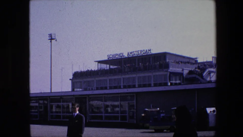 1967 8mm view of the Schiphol Amsterdam terminal with a suited man standing near the curb, a dark foreground passerby, visible observation deck visitors, and airport vehicles in front of the building.