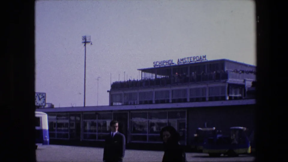 1967 8mm frame of the Schiphol Amsterdam airport frontage, showing the rooftop sign, observation deck visitors, small vehicles, a suited man, and a woman walking across the foreground.