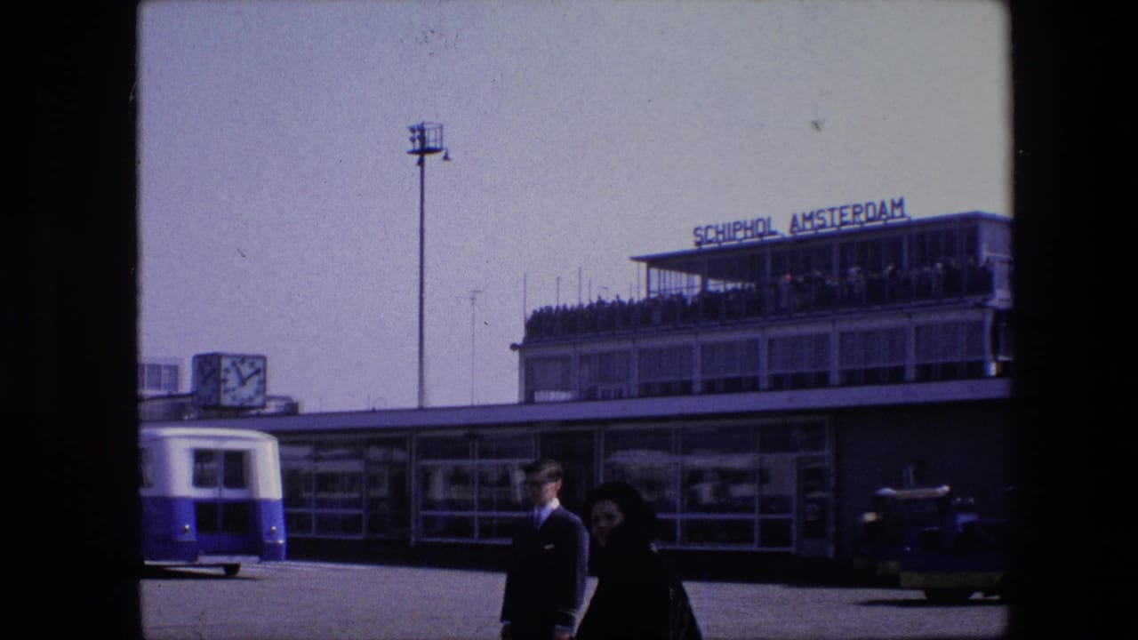 1967 8mm frame of Schiphol Airport in Amsterdam, with terminal signage, an airport bus, a large clock, and passengers walking across the tarmac.