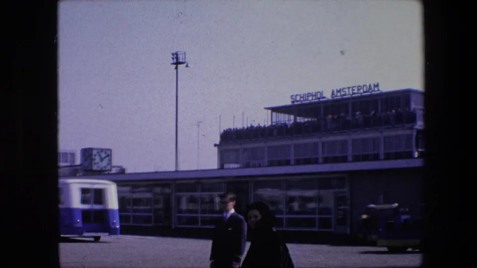 1967 8mm frame of Schiphol Airport in Amsterdam, with terminal signage, an airport bus, a large clock, and passengers walking across the tarmac.