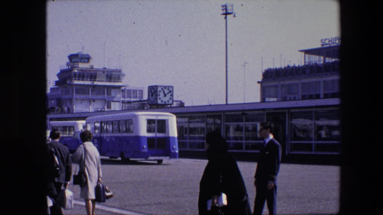 1967 Amsterdam Schiphol airport frame showing passengers in period clothing walking with bags near the terminal, control tower, clock, and parked airport bus.