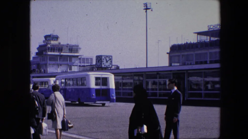 1967 Amsterdam Schiphol airport frame showing passengers in period clothing walking with bags near the terminal, control tower, clock, and parked airport bus.