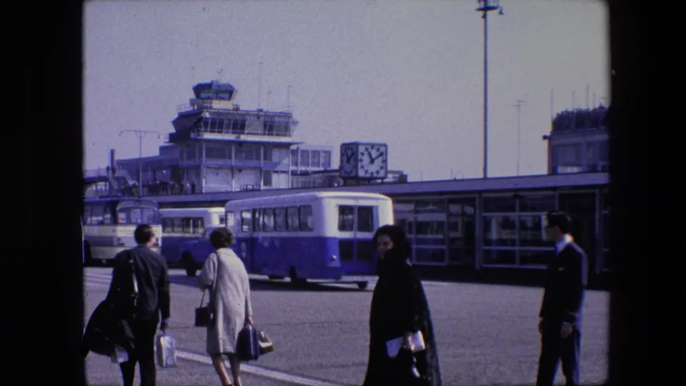 1967 8mm frame at Schiphol Airport, showing the control tower, terminal clock, blue-and-white bus, and several travelers moving across the apron with bags.