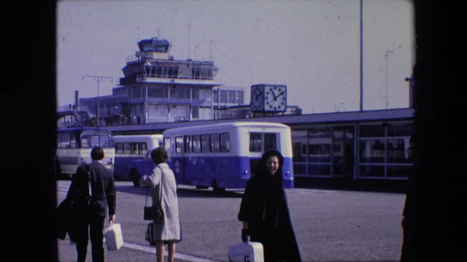 1967 Amsterdam Schiphol frame with a woman in a black coat carrying a suitcase in the foreground, while other travelers and airport buses stand near the terminal and control tower.