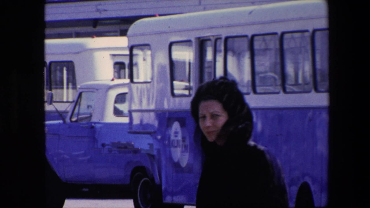 A woman in a dark fur-like coat moves through a daylight street scene near a blue-and-white bus marked with KLM lettering, with a blue truck or van also visible.