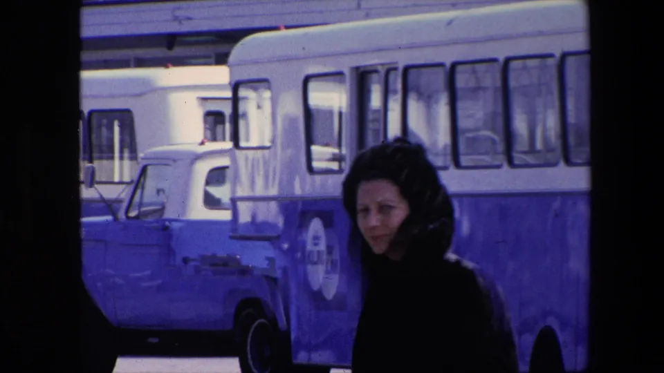 A woman in a dark fur-like coat moves through a daylight street scene near a blue-and-white bus marked with KLM lettering, with a blue truck or van also visible.