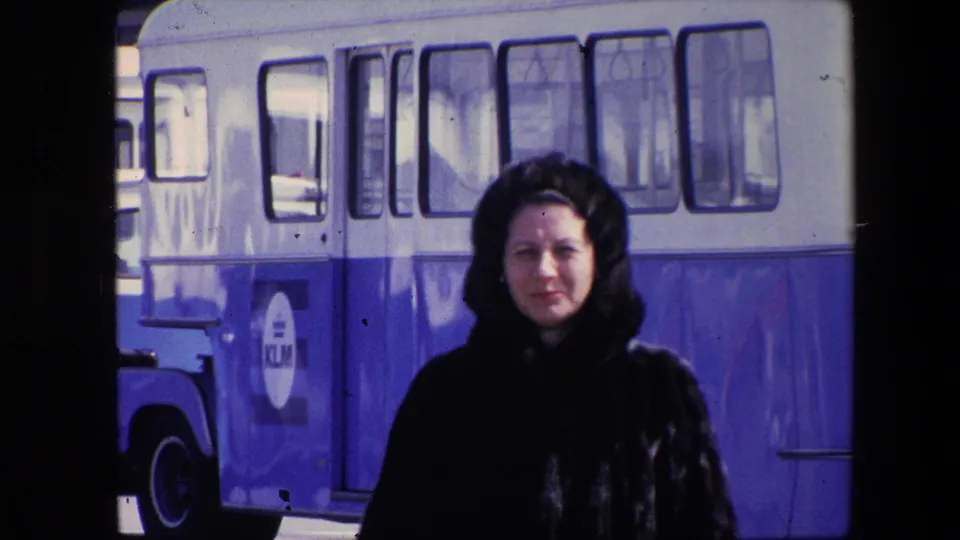 The woman stands or walks centered in the frame before a blue-and-white bus, with KLM lettering partly visible on the vehicle side.