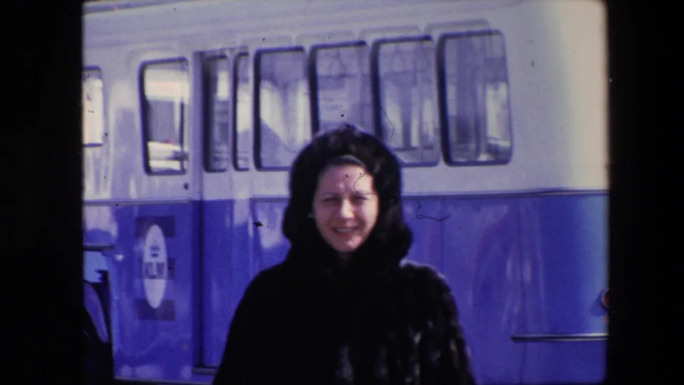 The woman appears to smile or speak toward the camera while standing near the blue-and-white KLM-marked bus in a faded 8mm frame.