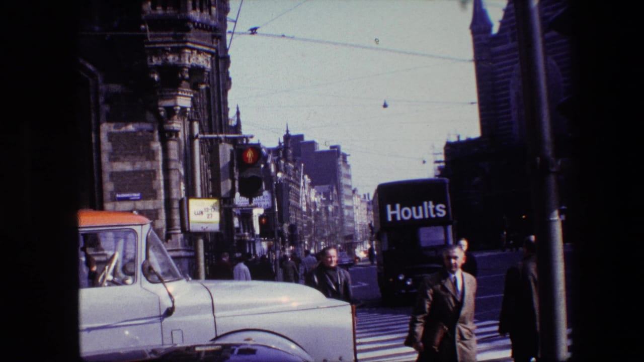 1967 Amsterdam street view with pedestrians at a zebra crossing, a white vintage truck, a black Hoults van, traffic lights, tram wires, and ornate buildings.