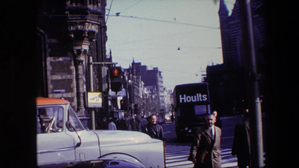 1967 Amsterdam street view with pedestrians at a zebra crossing, a white vintage truck, a black Hoults van, traffic lights, tram wires, and ornate buildings.
