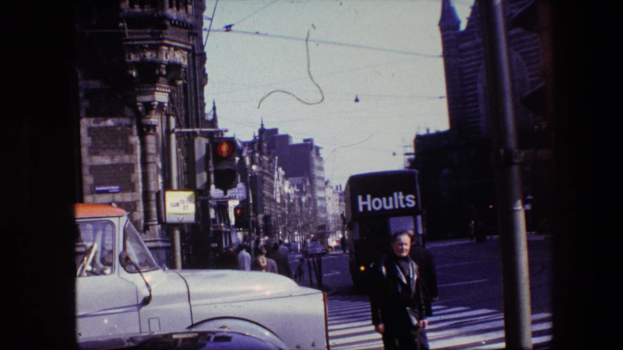 1967 Amsterdam street frame showing a man near the crosswalk, a white truck at left, a Hoults delivery van at right, overhead wires, and dense city architecture.