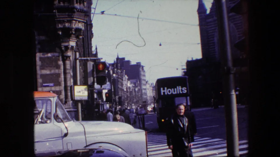 1967 Amsterdam street frame showing a man near the crosswalk, a white truck at left, a Hoults delivery van at right, overhead wires, and dense city architecture.