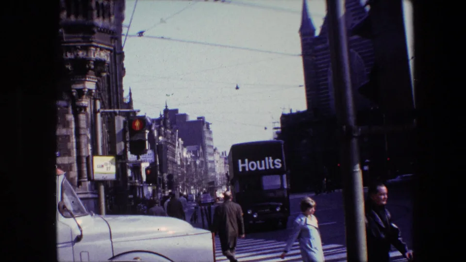 1967 Amsterdam street scene with pedestrians crossing in front of vintage vehicles, a Hoults van, traffic lights, tram wires, and tall urban buildings.
