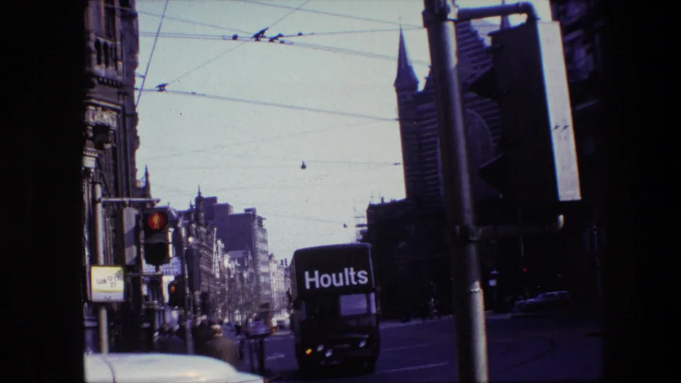 1967 Amsterdam street view with a Hoults van moving through the roadway, pedestrians in the distance, a traffic signal, overhead tram wires, and a prominent church-like spire.