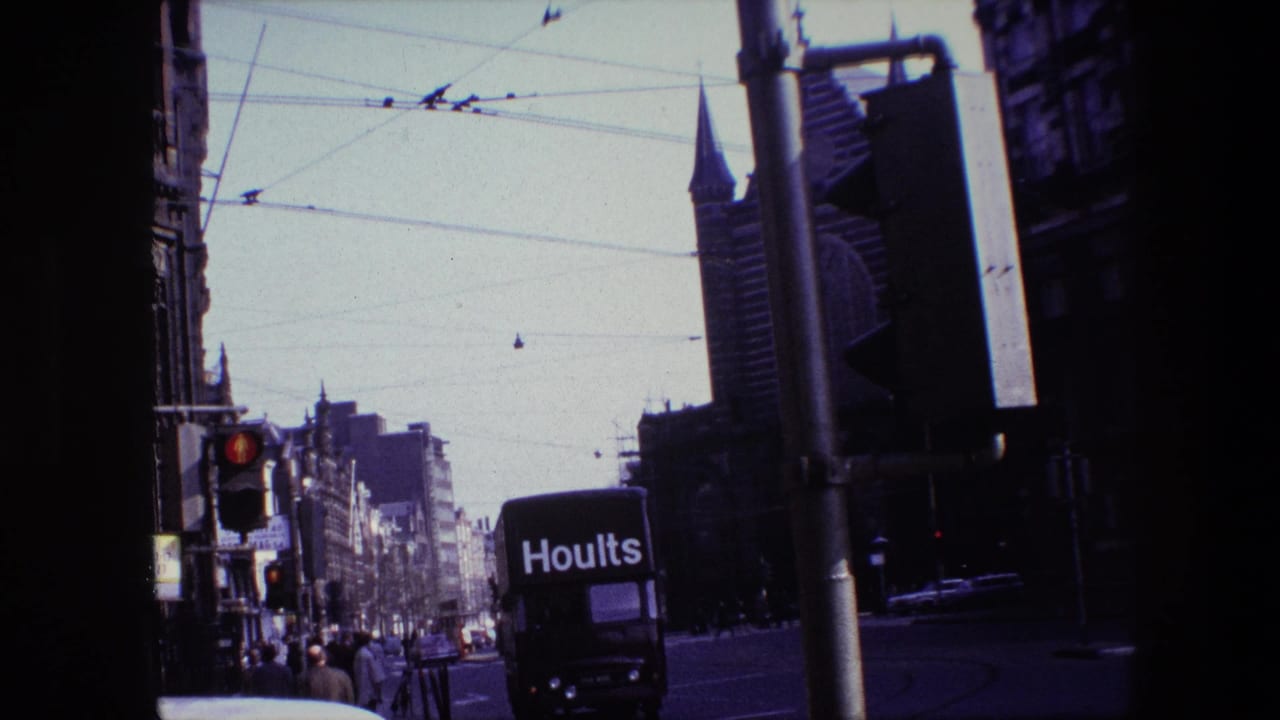 Amsterdam street scene from 1967 with a black Hoults truck in traffic beneath overhead tram wires, pedestrians along the sidewalk, and a spire rising beyond the buildings.