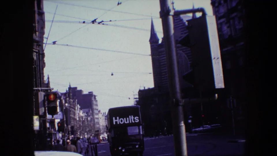 Amsterdam street scene from 1967 with a black Hoults truck in traffic beneath overhead tram wires, pedestrians along the sidewalk, and a spire rising beyond the buildings.