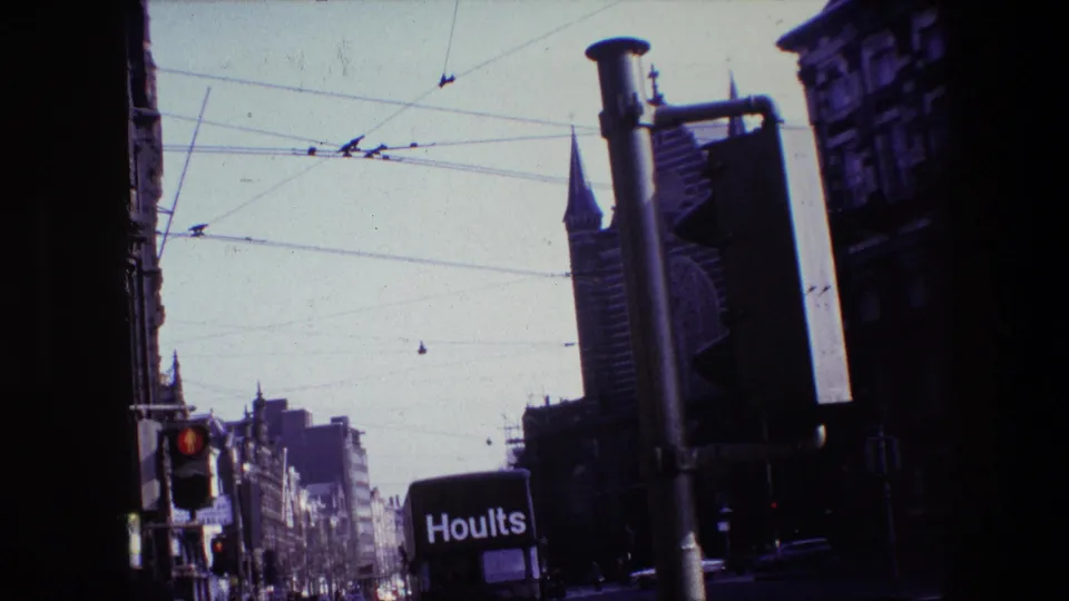 Angled 1967 Amsterdam street view showing the Hoults truck lower in frame, a red traffic light, dense overhead tram wires, and a pointed spire among city buildings.