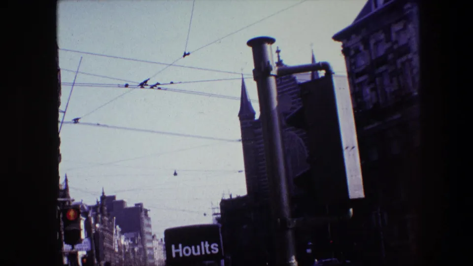 Vehicle-framed Amsterdam street footage from 1967 with overhead tram wires crossing the sky, pedestrians near the curb, a Hoults truck, and historic-looking architecture in the distance.