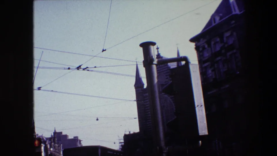 Late frame of the 1967 Amsterdam street view with tram wires overhead, a traffic signal at left, the Hoults truck partly visible, and a spire and older buildings in the background.