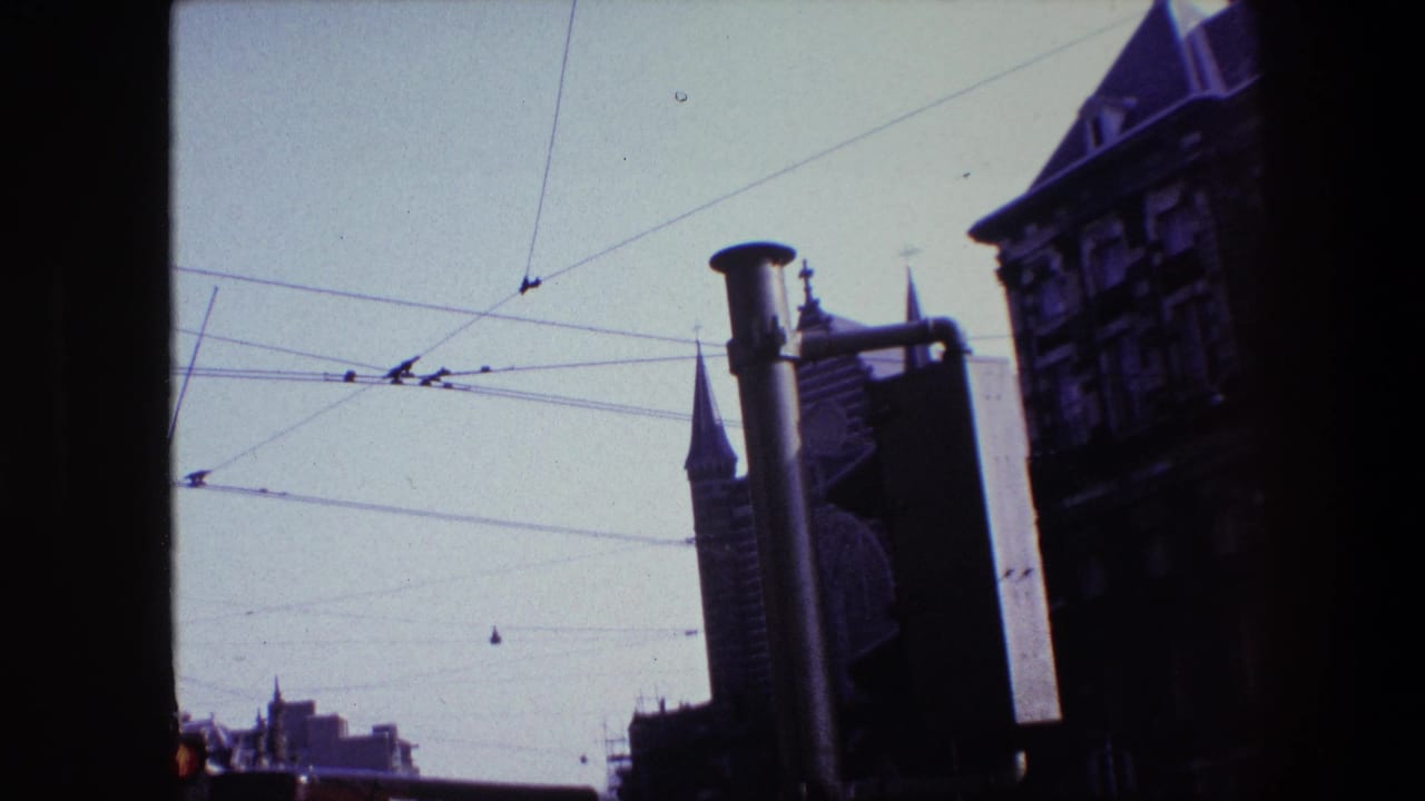 1967 Amsterdam home-movie frame showing church spires, overhead trolley wires, a street pole, and shadowed historic facades from a low moving viewpoint.