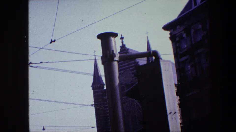 1967 Amsterdam home-movie still with a foreground street pole, intersecting overhead wires, church spires, and shadowed historic architecture.
