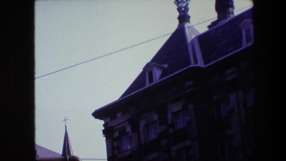 1967 Amsterdam home-movie frame emphasizing a dark ornate roofline with dormers, nearby church spires, crosses, and a diagonal overhead wire.