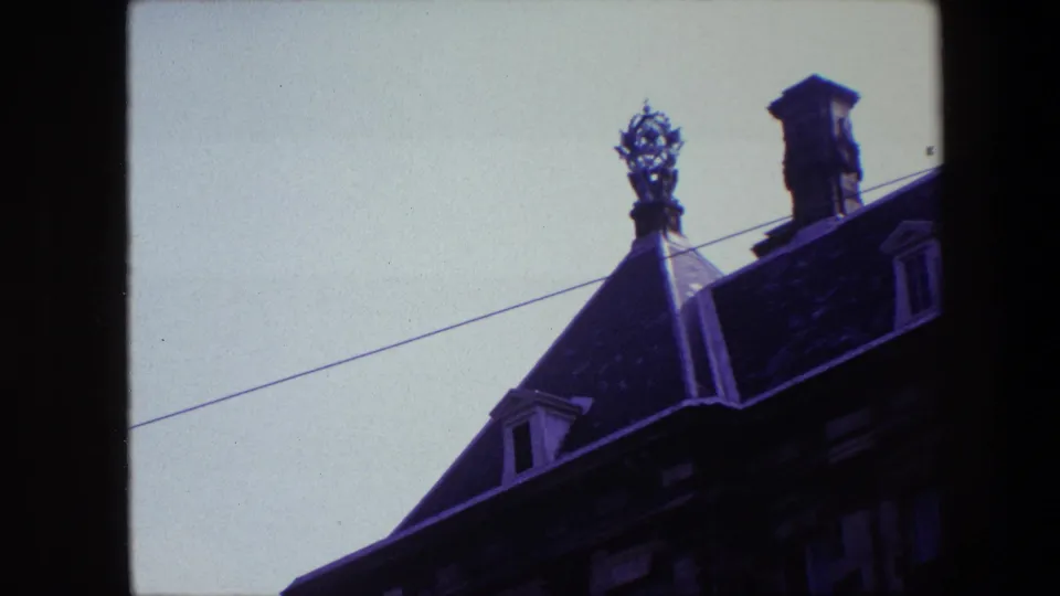 1967 Amsterdam frame showing the upper portion of a steep old roof with a decorative rooftop ornament, chimney, dormer, and overhead wire.