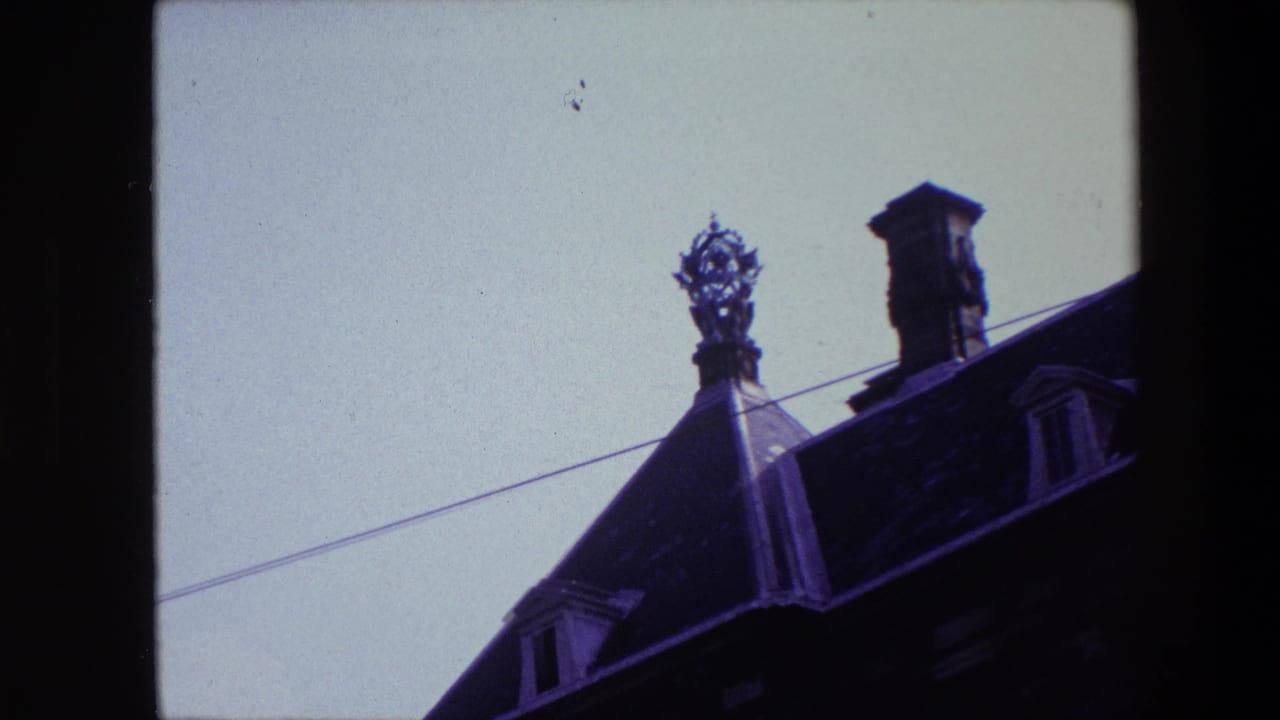 1967 archival 8mm frame associated with Amsterdam showing an ornate rooftop finial or grotesque near a chimney and dormer, silhouetted against a pale sky.