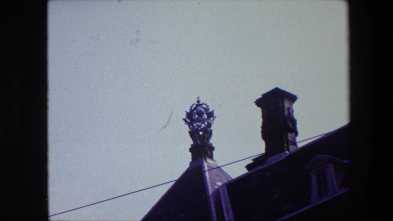 1967 archival 8mm frame associated with Amsterdam showing a decorative rooftop figure and chimney on a dark historic roof beneath open sky.