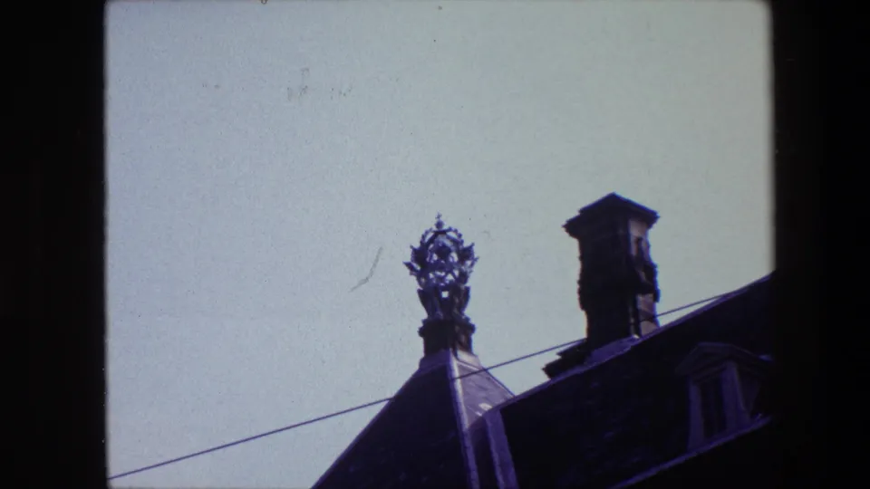 1967 archival 8mm frame associated with Amsterdam showing a decorative rooftop figure and chimney on a dark historic roof beneath open sky.