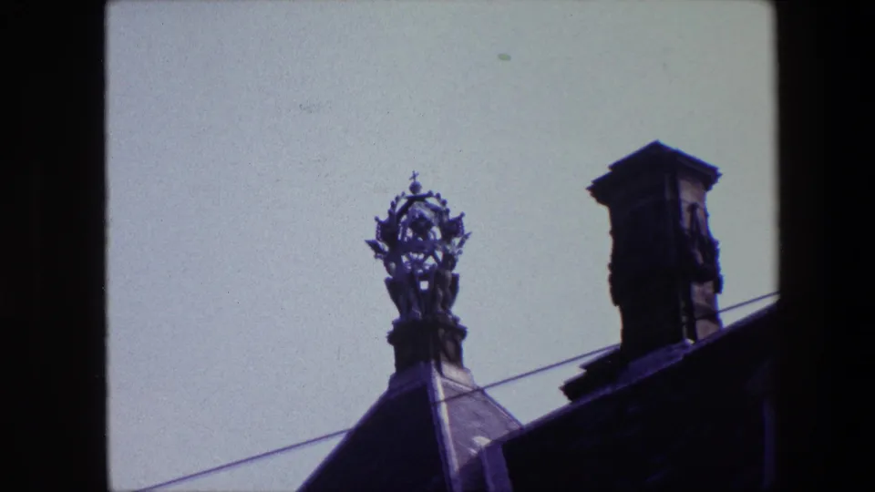 1967 archival 8mm frame associated with Amsterdam showing a closer view of an ornate rooftop sculpture, with a chimney, roof slope, and diagonal wire visible.