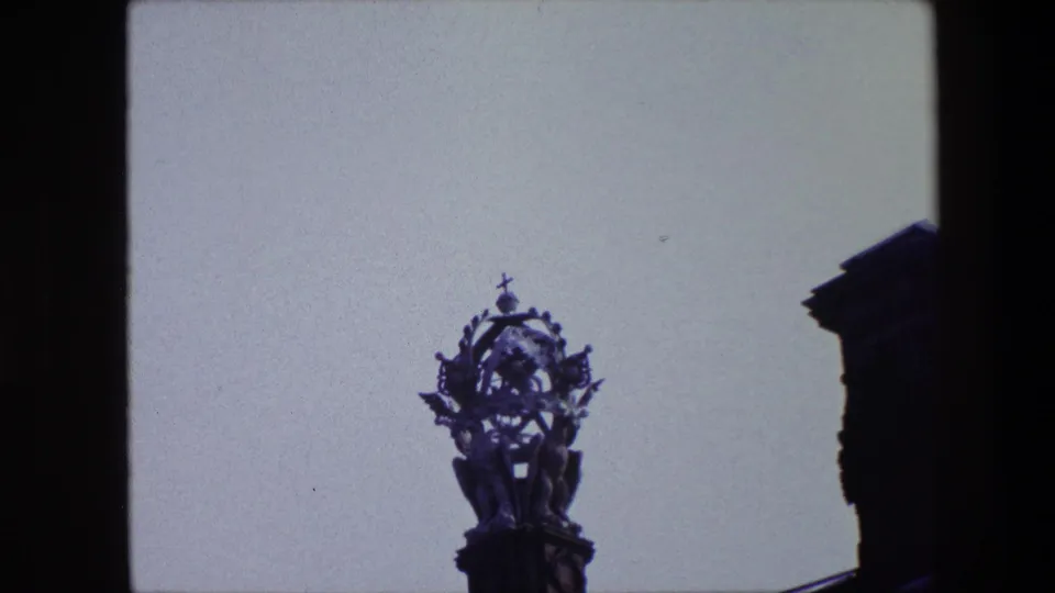 1967 archival 8mm frame associated with Amsterdam showing the ornate rooftop figure isolated against a pale sky, with part of the building edge cropped at right.
