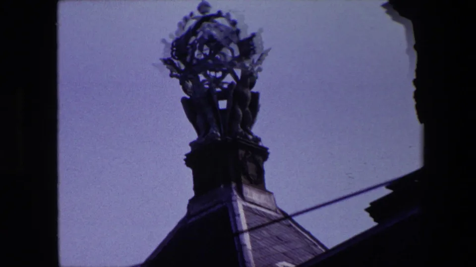 1967 Amsterdam home-movie frame of the church steeple above a roofline, with dark architectural framing and overhead street wires crossing below.