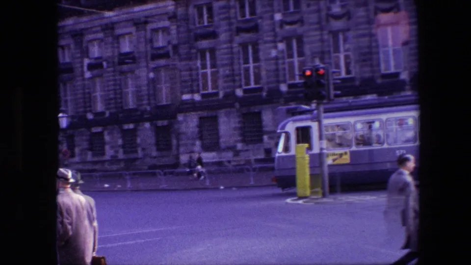 1967 Amsterdam street scene with masonry facades, overhead wires, a tram or bus, a small van, and pedestrians crossing or standing near the road.